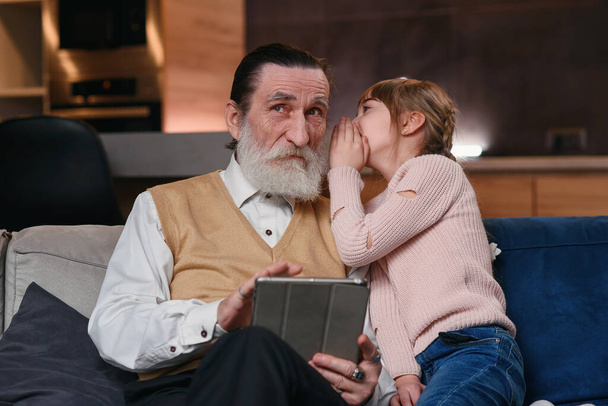 Slow motion of cute joyful 12-aged girl with funny pigtails which whispering on granddads ear her secret while they sitting together on the sofa at home - Photo, Image