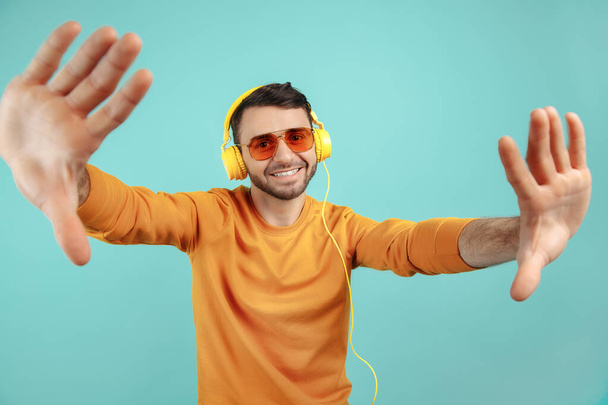 Careless bearded young man wearing sunglasses listening to music and dancing on cyan background. - Fotó, kép