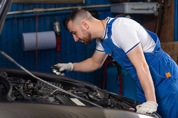 Handsome young male auto mechanic in special uniform clothes holding a flashlight, looking for breakdown and repairing under the hood in the car engine in a car workshop - Φωτογραφία, εικόνα