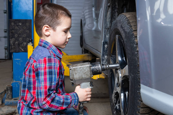 A young boy, a young auto worker, makes a tire change with a pneumatic wrench in the garage of a service station. A child learns the mechanics changing profession in auto repair service. - Foto, immagini