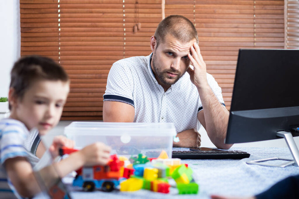 Egy fiatal férfi apa sajnálatos módon megpróbál egy számítógépnél dolgozni egy távoli munkahelyen az elszigeteltség időszakában a coronovírus világjárvánnyal kapcsolatban, lágy fókuszban. Otthoni munka kisgyermekekkel - Fotó, kép