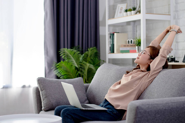Mulher asiática desfrutando e relaxando depois de usar o computador Ela está sentada no sofá em casa
. - Foto, Imagem