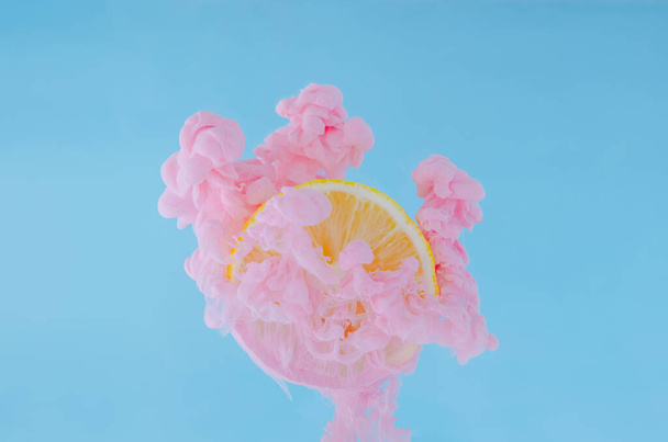 Φέτα λεμόνι με μερική εστίαση της διάλυσης ροζ αφίσα χρώμα στο νερό σε μπλε φόντο για το καλοκαίρι, αφηρημένη και φόντο έννοια. - Φωτογραφία, εικόνα