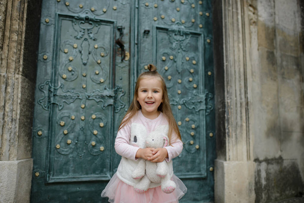 Klein meisje houdt een speelgoedkonijntje in haar handen en glimlacht. Een close-up. Meisje poserend op de achtergrond van de deuren van de oude kerk. - Foto, afbeelding