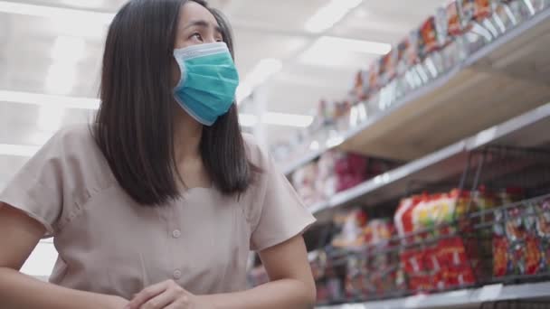 Asian woman in protective mask walking with shopping cart at grocery store during covid-19 pandemic crisis, scared worry anxiety concern, new normal lifestyle, after covid, grocery store aisle - Footage, Video