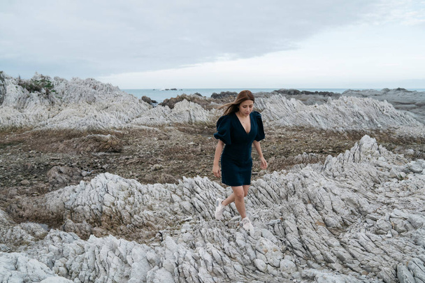 海の近くの石の上に若いアジアの女性の肖像画。ニュージーランド・カイコラでアウトドアレクリエーションを楽しむ美しい女の子が海岸に休んでいます. - 写真・画像