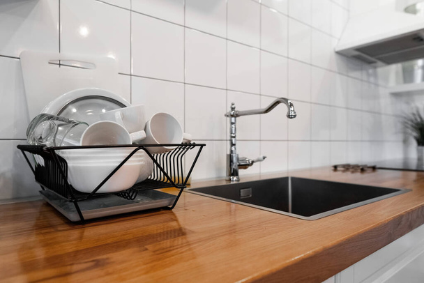 Budget and lightweight antimicrobial dish drainer with drain board at modern scandinavian kitchen. Dish rack holds many dishes and cups against wooden countertop, white wall tiles, sink and faucet - Photo, Image