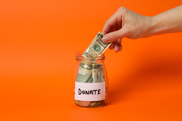 Vrouwelijke hand zet geld in glazen pot op oranje achtergrond - Foto, afbeelding