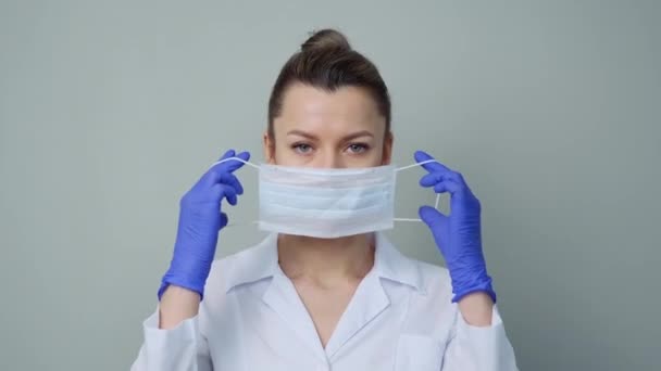 A young woman doctor in a white robe and gloves puts on a protective mask on her face. - Πλάνα, βίντεο