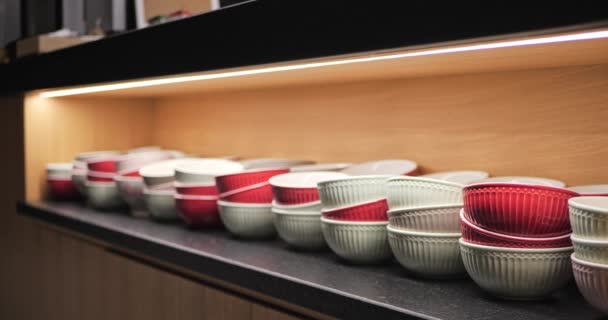 Red and white ceramic bowls in a wooden kitchen cabinet. Focus transition. Tableware lighted from above in a cupboard, modern interior showcase. - Footage, Video