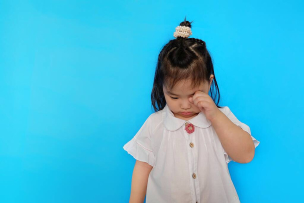 Eine süße asiatische Kleinkindfrau ist aufgebracht, weint und wischt sich mit der Hand Tränen aus den Augen. Einfacher blauer Hintergrund. - Foto, Bild