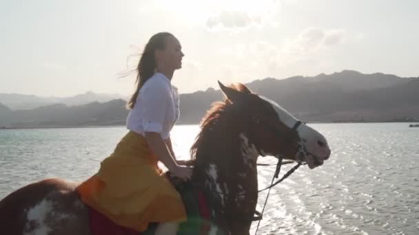 Valkoihoinen blondi tyttö ratsastus hevonen läpi veden auringonlaskun hidastettuna
 - Materiaali, video