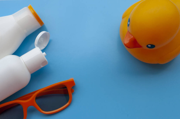Sonnencreme, orangefarbene Sonnenbrille, Muscheln, orangefarbene Plastikente auf hellblauem Hintergrund. Konzept von Sommer, Urlaub, Erholung, Meer. - Foto, Bild