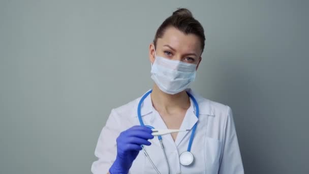A female medical worker in a white robe and mask holds and looks at an electronic thermometer. - Video, Çekim
