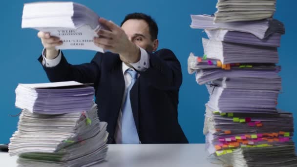 Un oficinista de mediana edad cambia montones de papeles en su escritorio. Fondo azul aislado
. - Imágenes, Vídeo