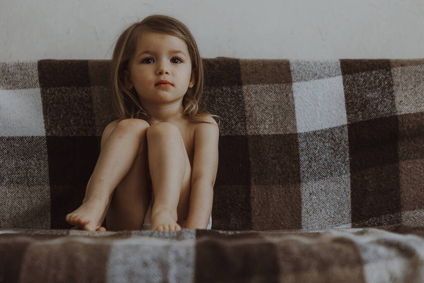 cute little girl dressed in panties sits on the couch in her parents' room hugging her knees and sadly looking ahead - 写真・画像