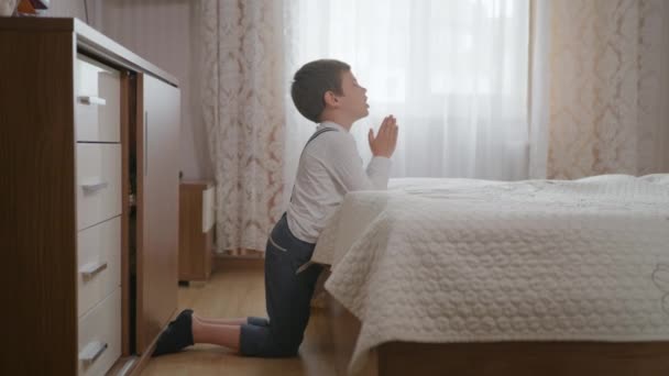 religion, little Christian boy with faith and hope in heart with closed eyes clasped hands prays to God on his knees in room near bed - Кадры, видео