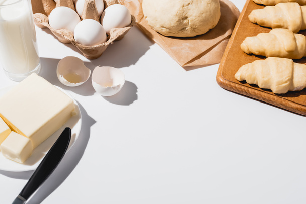 verse croissants op houten snijplank bij rauw deeg, melk, boter en eieren op witte achtergrond - Foto, afbeelding