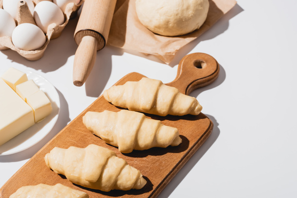 fresh croissants on wooden cutting board near raw dough, rolling pin, butter and eggs on white background - Photo, Image