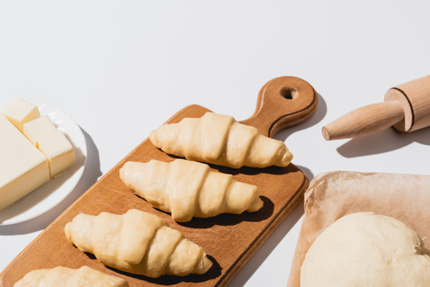fresh croissants on wooden cutting board near raw dough, rolling pin, butter on white background - Fotografie, Obrázek
