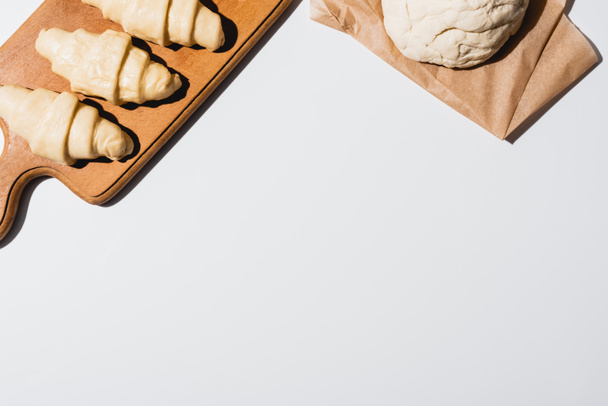 top view of fresh croissants on wooden cutting board near raw dough on white background - Photo, Image