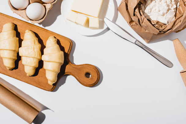 bovenaanzicht van rauwe croissants op houten snijplank bij ingrediënten op witte achtergrond - Foto, afbeelding