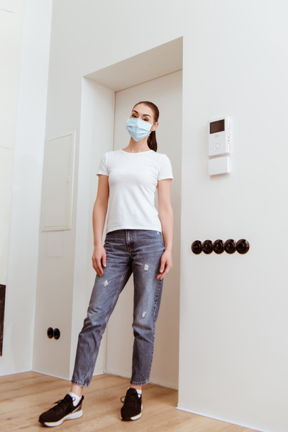 Woman in medical mask looking at camera while standing in hallway at home  - Foto, immagini
