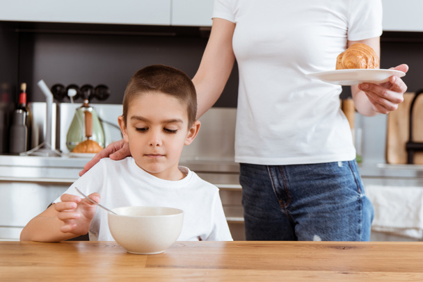 Mother holding croissant near son eating breakfast in kitchen  - Фото, зображення