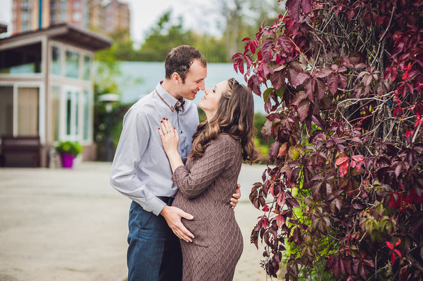 A pregnant young woman and her husband. A happy family standing at the red autumn hedge, holding belly. pregnant woman relaxing in the park. - Photo, image