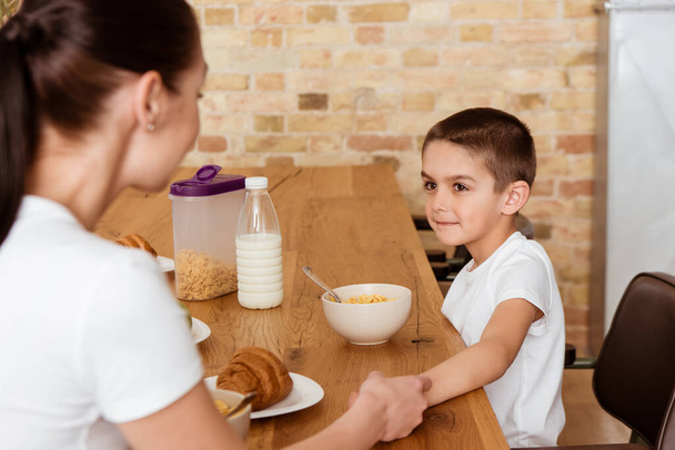 Selective focus of boy and mother holding hands near cereals and croissants on kitchen table  - 写真・画像