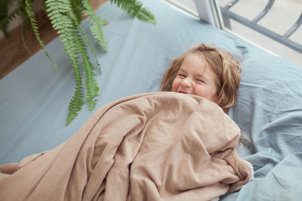 Menina alegre feliz encontra-se em um sofá azul perto da janela, a criança sorri no quarto em casa, interior
 - Foto, Imagem