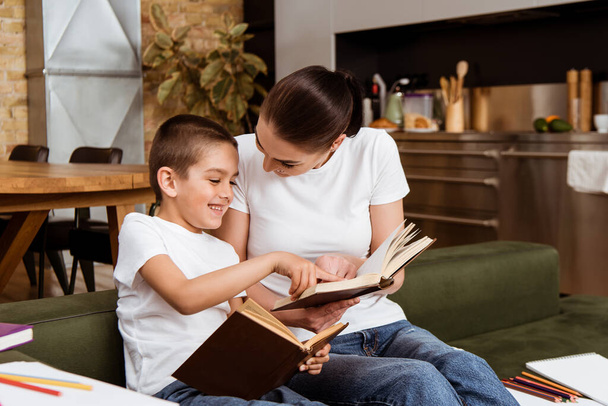 Selective focus of smiling boy pointing with finger while reading books with mother on couch  - Foto, Bild
