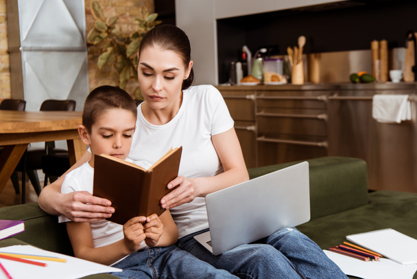 Enfoque selectivo del libro de lectura de madre e hijo cerca de la computadora portátil, papel y lápices de color en el sofá
  - Foto, Imagen