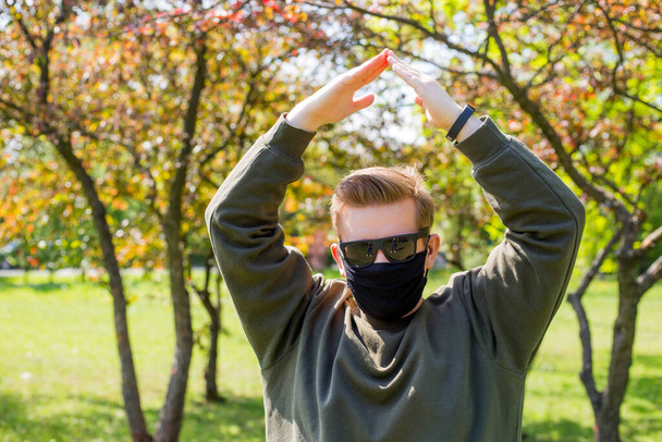 Άντρας με μια προστατευτική μαύρη μάσκα που κάνει σήμα στο σπίτι σε ένα πάρκο της πόλης. Αντιγραφή χώρου. Αντίληψη σύμβολο σπίτι γίνεται με τα χέρια στο φόντο της φύσης. Guy κτίριο κατασκευή έννοια κρατήσει στέγη πάνω από τα κεφάλια του - Φωτογραφία, εικόνα