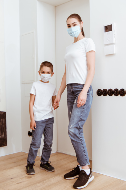 Mother and son in medical masks holding hands in hallway  - 写真・画像