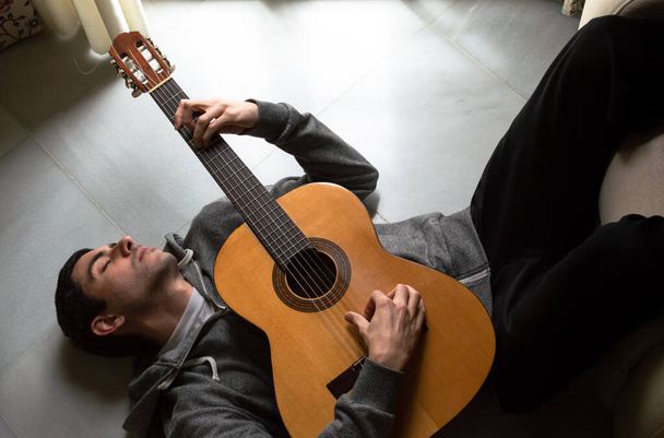 Homem relaxado deitado no chão tocando guitarra perto de uma janela brilhante em casa. Guitarrista apreciando a música, momento harmonioso
. - Foto, Imagem