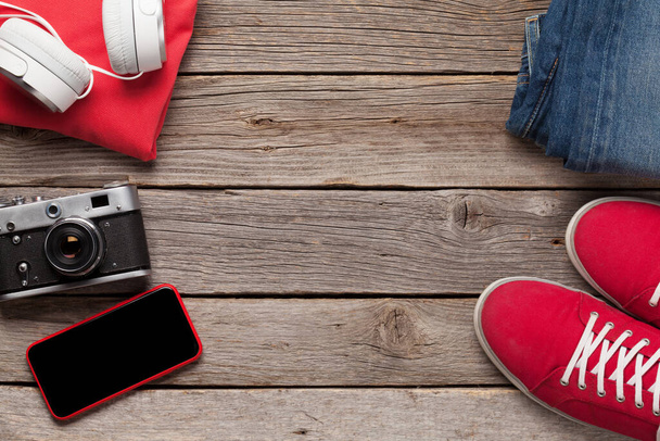 Kleding en accessoires. Sneakers, jeans, koptelefoon en smartphone. Stedelijke outfit voor alledaagse of reis vakantie op houten achtergrond met kopieerruimte. Bovenaanzicht vlak lay - Foto, afbeelding