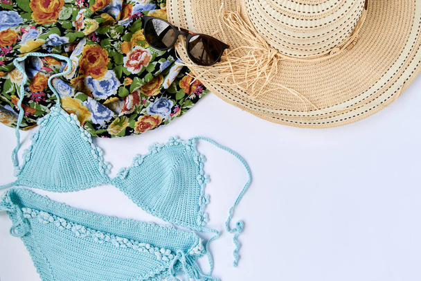 Flat lay picture of summer vacation beach accessories, isolated on white background. Mint light blue swimsuit, sunglasses, colorful flowery scarf and straw hat. Tropical vacation planning. - Photo, Image