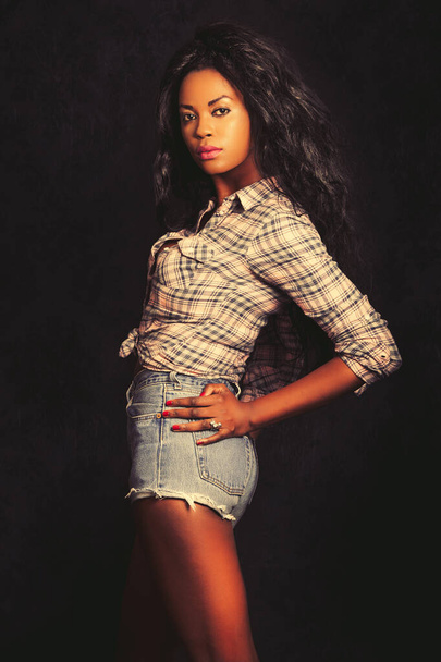 Schöne junge schwarze Frau. Afro Modell mit dunklem Hintergrund. Lässig gekleidet mit kurzen Jeans und Hemd. Schwarze lange Haare. Posieren im Studio, sinnliche und modellierte Schönheit. - Foto, Bild