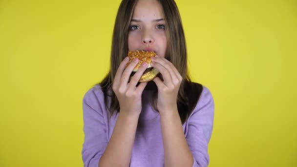 Tiener meisje in een roze trui eet een hamburger op gele achtergrond met kopieerruimte - Video