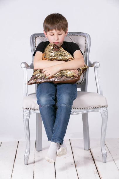 Λυπηρό αγόρι που κάθεται σε μια καρέκλα αγκαλιάζοντας ένα μαξιλάρι. - Φωτογραφία, εικόνα