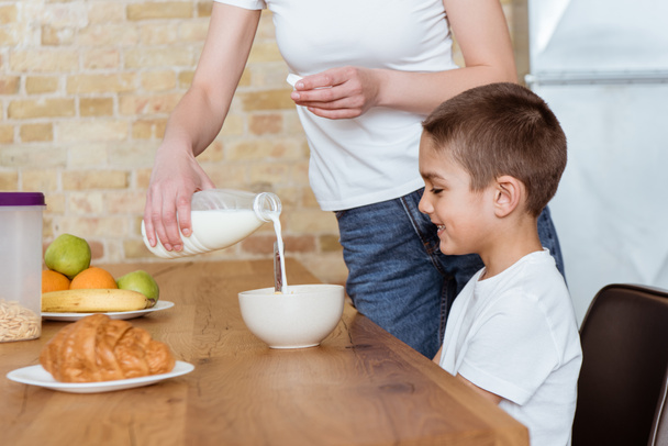 Selective focus of mother pouring milk in bowl with cereals near smiling son in kitchen  - Foto, Imagem