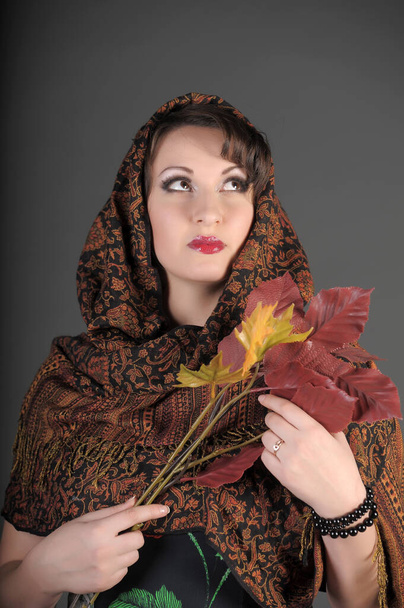 beautiful Russian brunette woman with a scarf on her head and autumn leaves in her hands, photo in the studio on a gray background - Photo, image
