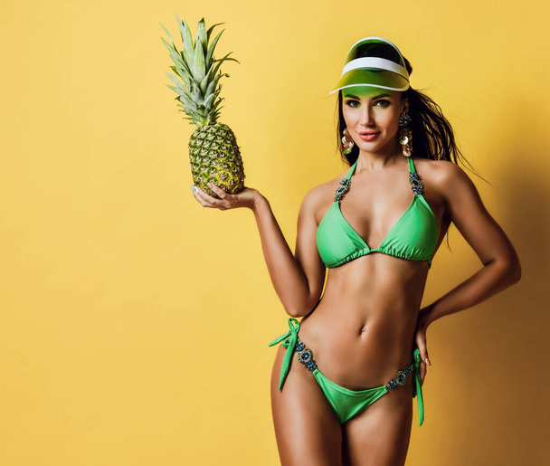 Young tanned slim brunette woman in sexy green bikini with decorations and visor is posing holding fresh pineapple - Foto, Imagen