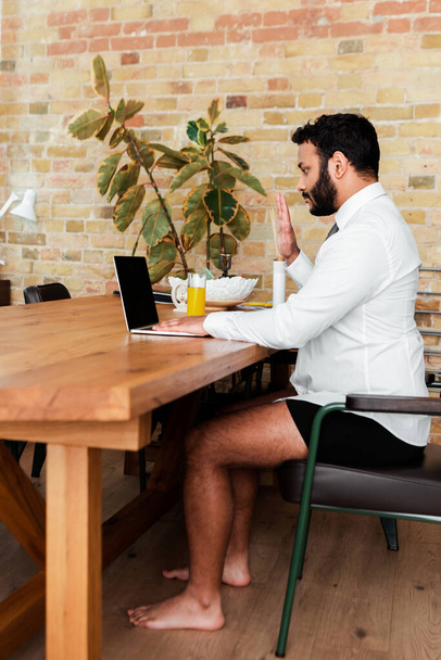 бородатий афроамериканський чоловік у сорочці та нижній білизні сидить біля ноутбука та махає рукою, маючи відеодзвінок
  - Фото, зображення