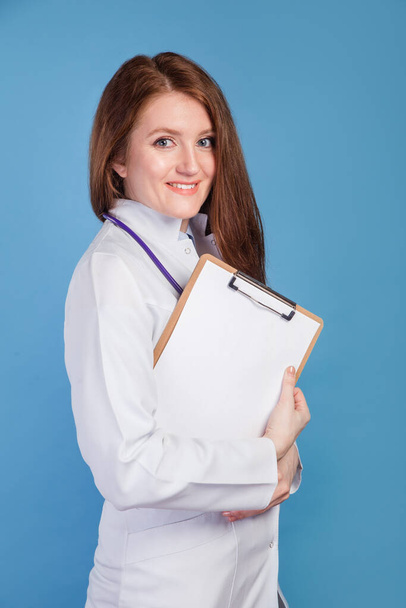 Young happy smiling woman doctor holding folder with diagnosis on blue background - 写真・画像