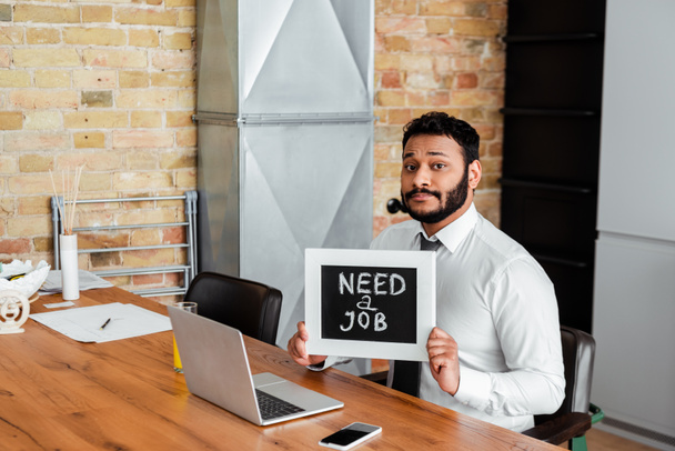 bearded african american man holding chalkboard with need a job lettering near gadgets  - Photo, Image