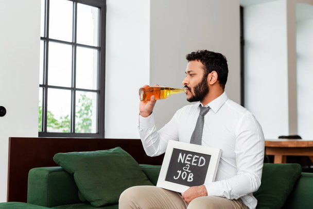 werkloze Afro-Amerikaanse man drinkt bier en houdt schoolbord met behoefte aan een baan belettering thuis  - Foto, afbeelding