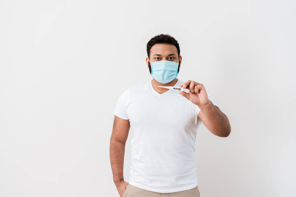 hombre afroamericano enfermo en máscara médica de pie con la mano en el bolsillo y la celebración de termómetro digital cerca de la pared blanca
  - Foto, imagen