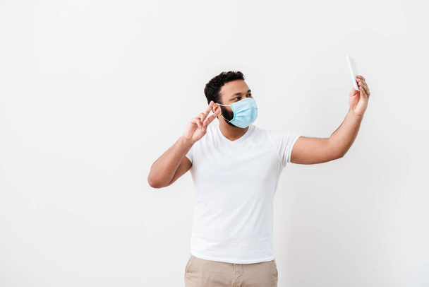 african american man in medical mask showing peace sign and taking selfie near white wall  - Foto, imagen